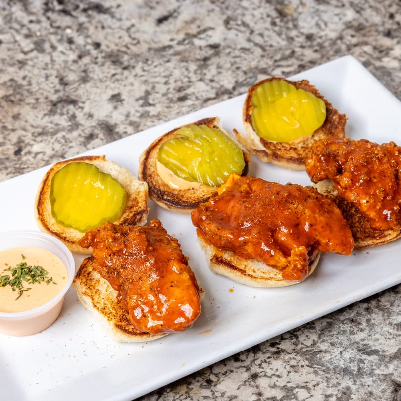 Spicy chicken sliders with pickles and a side of dipping sauce on a white plate.