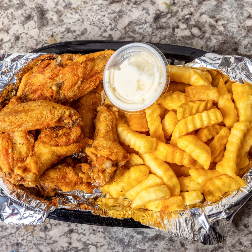 Crispy fried chicken wings and crinkle-cut fries with a side of dipping sauce in a foil-lined basket.