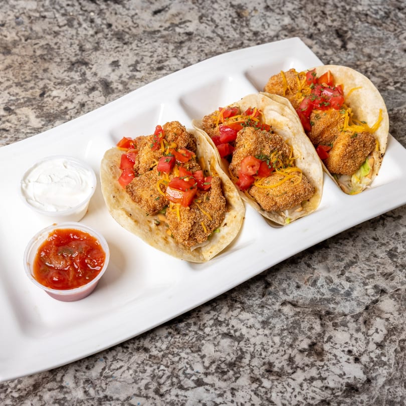 Three crispy chicken tacos topped with diced tomatoes, shredded cheese, and herbs, served with salsa and sour cream.