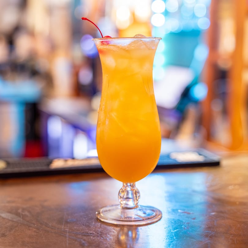 A vibrant orange cocktail with ice and a cherry garnish sits on a bar top at DaBomb Sports Grill.